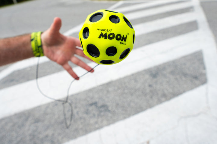Hand holding a yellow and black Waboba Moon Bounceback ball on a white background